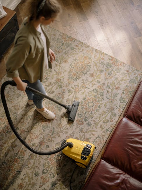 A woman using a yellow vacuum cleaner to deep clean a patterned area rug inside a living room with wooden flooring. She is wearing a beige jacket, dark pants, and sneakers, and is standing on the rug,