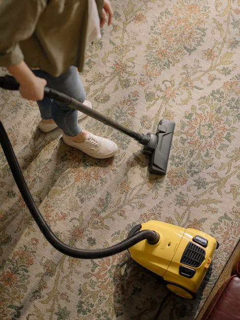 A woman using a yellow vacuum cleaner to deep clean a patterned area rug inside a living room with wooden flooring. She is wearing a beige jacket, dark pants, and sneakers, and is standing on the rug,