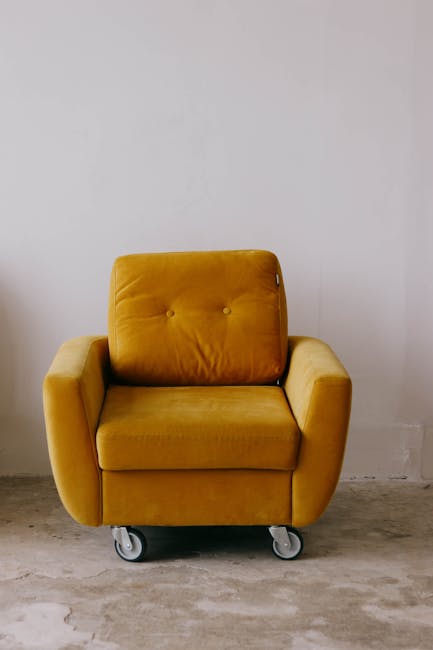 A close-up view of a living room corner featuring a light grey upholstered armchair with wooden legs positioned next to a window with white, semi-transparent curtains that allow soft natural light to